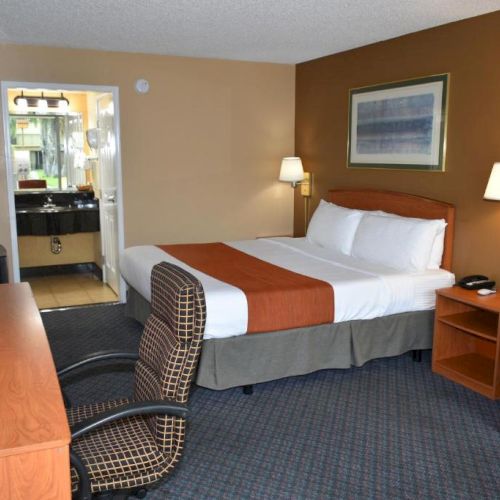 A hotel room with a bed, two bedside lamps, a desk and chair, a sofa, a small table, and an open door to a bathroom, all in warm tones.