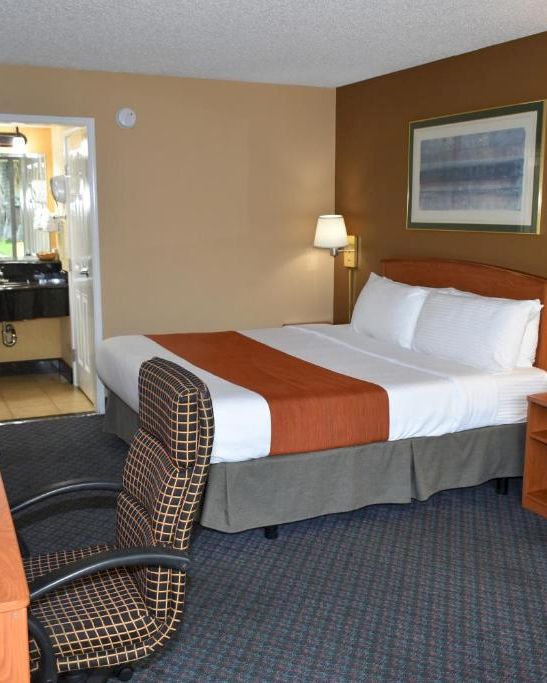 A hotel room with a bed, two bedside lamps, a desk and chair, a sofa, a small table, and an open door to a bathroom, all in warm tones.