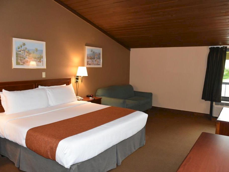 A hotel room with a large bed, white linens, brown runners, a sofa, a desk, wall art, and a window with dark curtains, under a wood-paneled ceiling.